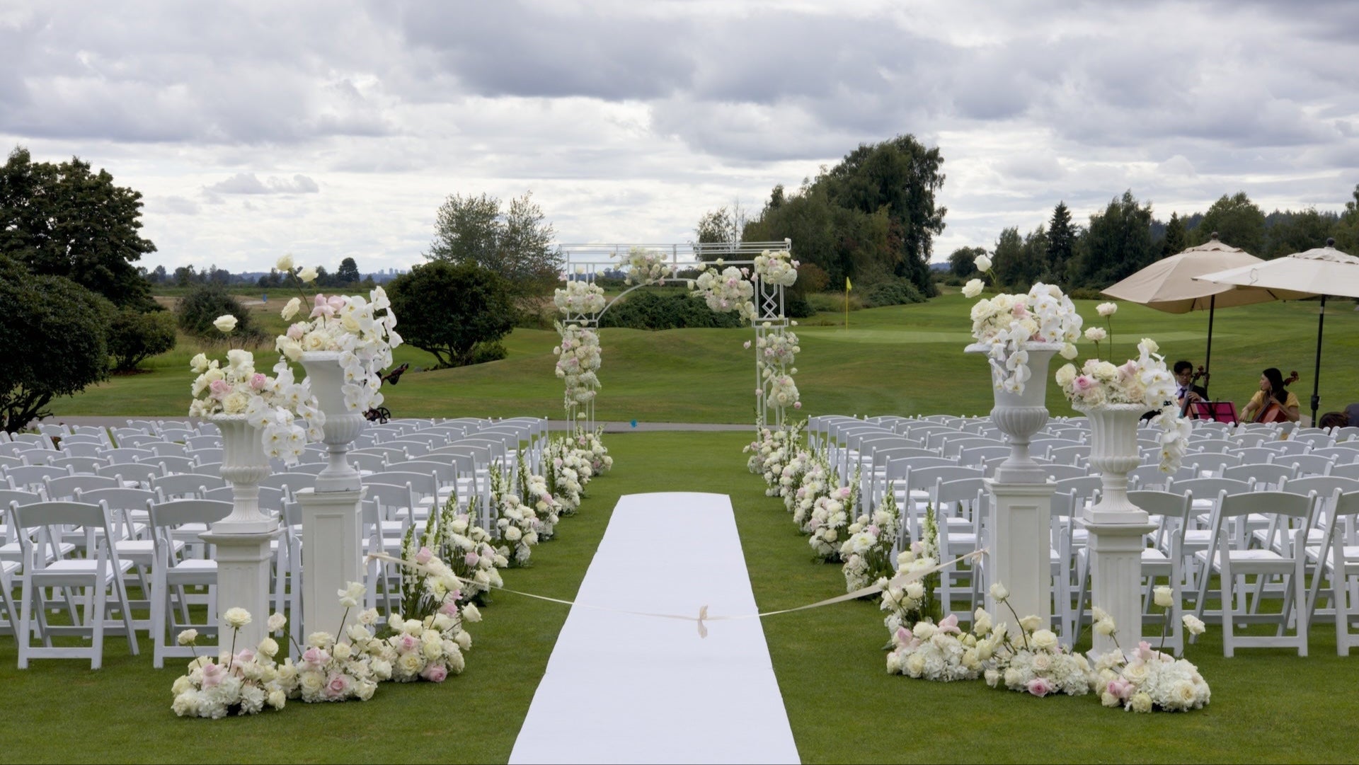 Wedding setup with white and light pink floral arch, floral aisle arrangements, statement arrangements, chairs, and a white aisle runner on Swaneset Bay Resort & Country Club grassy field.