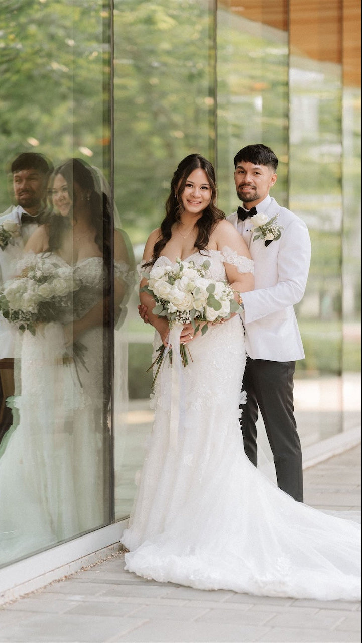 Couple in wedding attire standing in front of a glass wall with reflections.