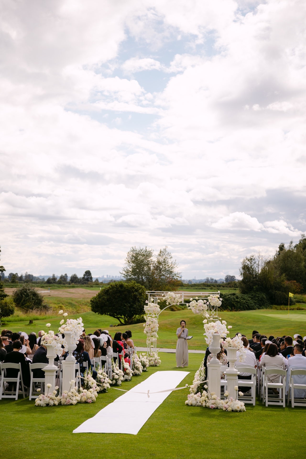 Wedding ceremony setup with floral arch and guests on a grassy field.