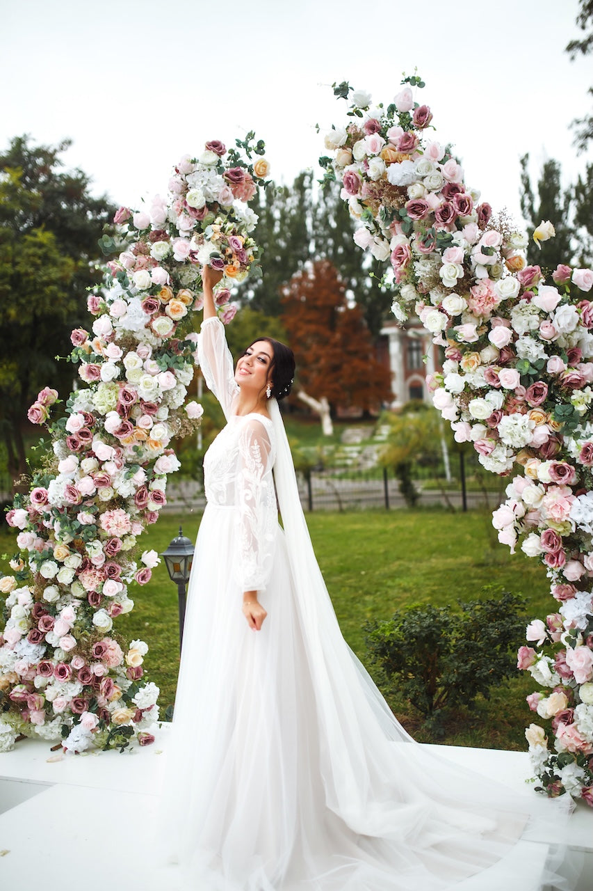 Bride in a white dress standing between two floral arches in an outdoor setting.