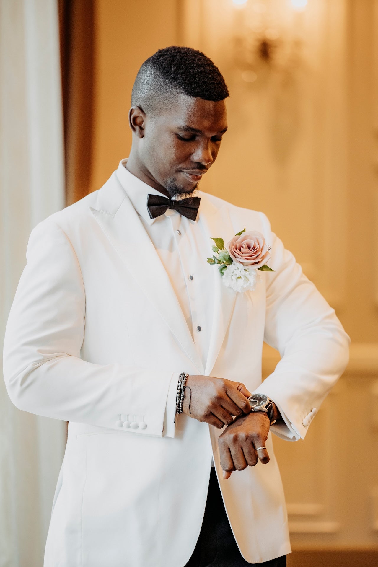 Groom in a white tuxedo with a bow tie and boutonniere, adjusting his watch.