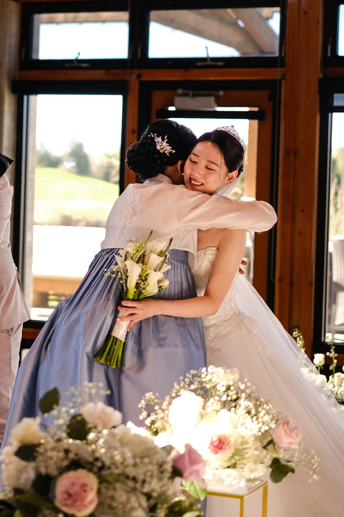 Two women embracing in a bright indoor setting with large windows and floral arrangements in the foreground. One wears a white bridal gown and veil, and the other wears a white top with a dusty blue skirt.
