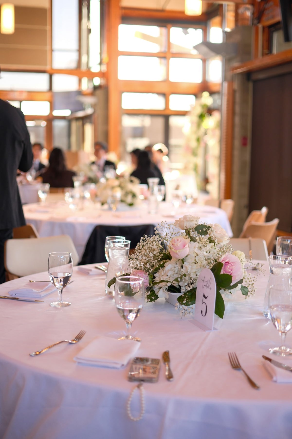 A round wedding table in Riverway Clubhouse Burnaby, set with a white tablecloth, clear wine glasses, silverware, a small floral centerpiece with pink roses and white flowers, and a vertical place card.