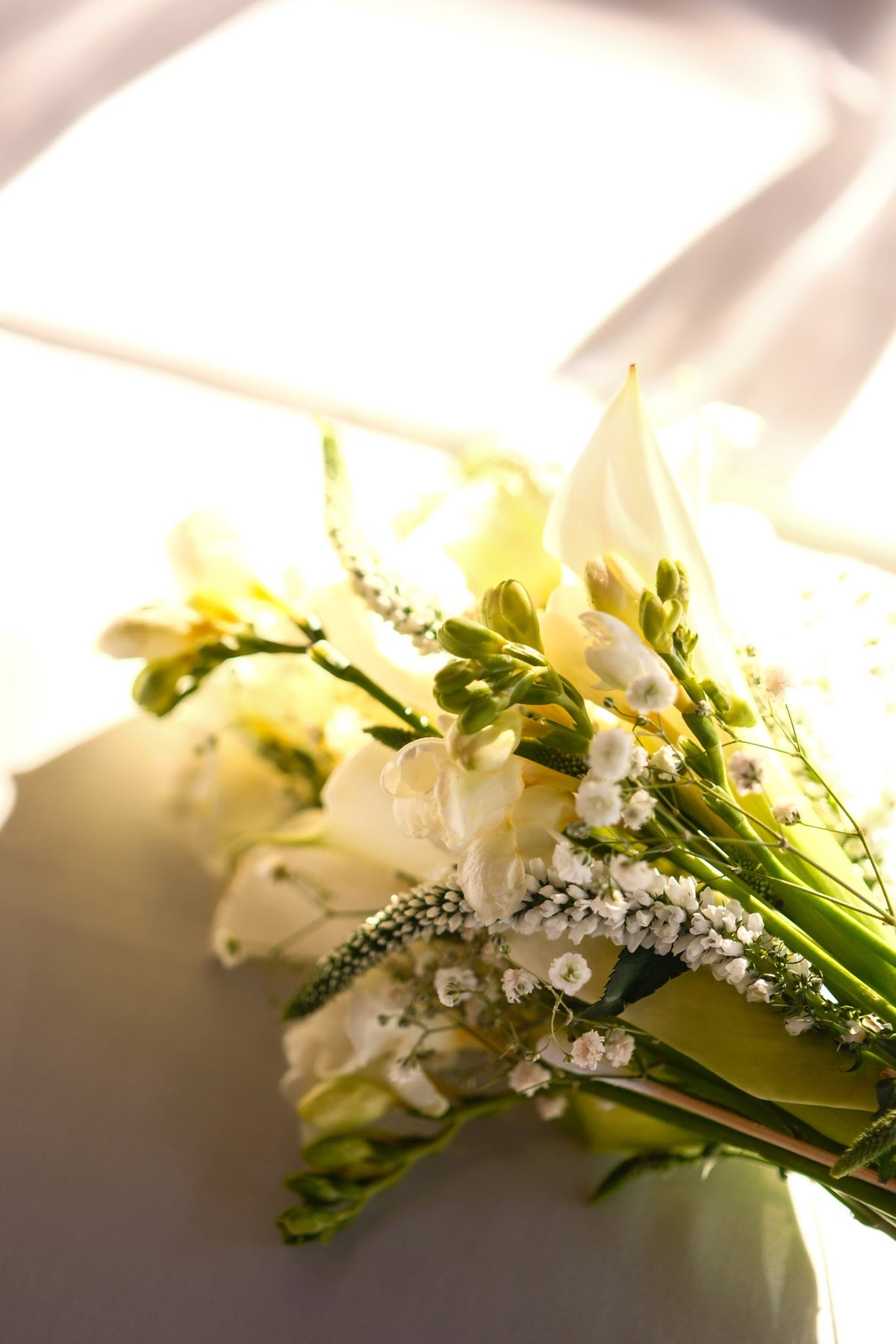 A close-up of a floral bouquet with white and off white flowers, green stems, and small white filler blossoms, photographed in bright natural light against a soft light background.