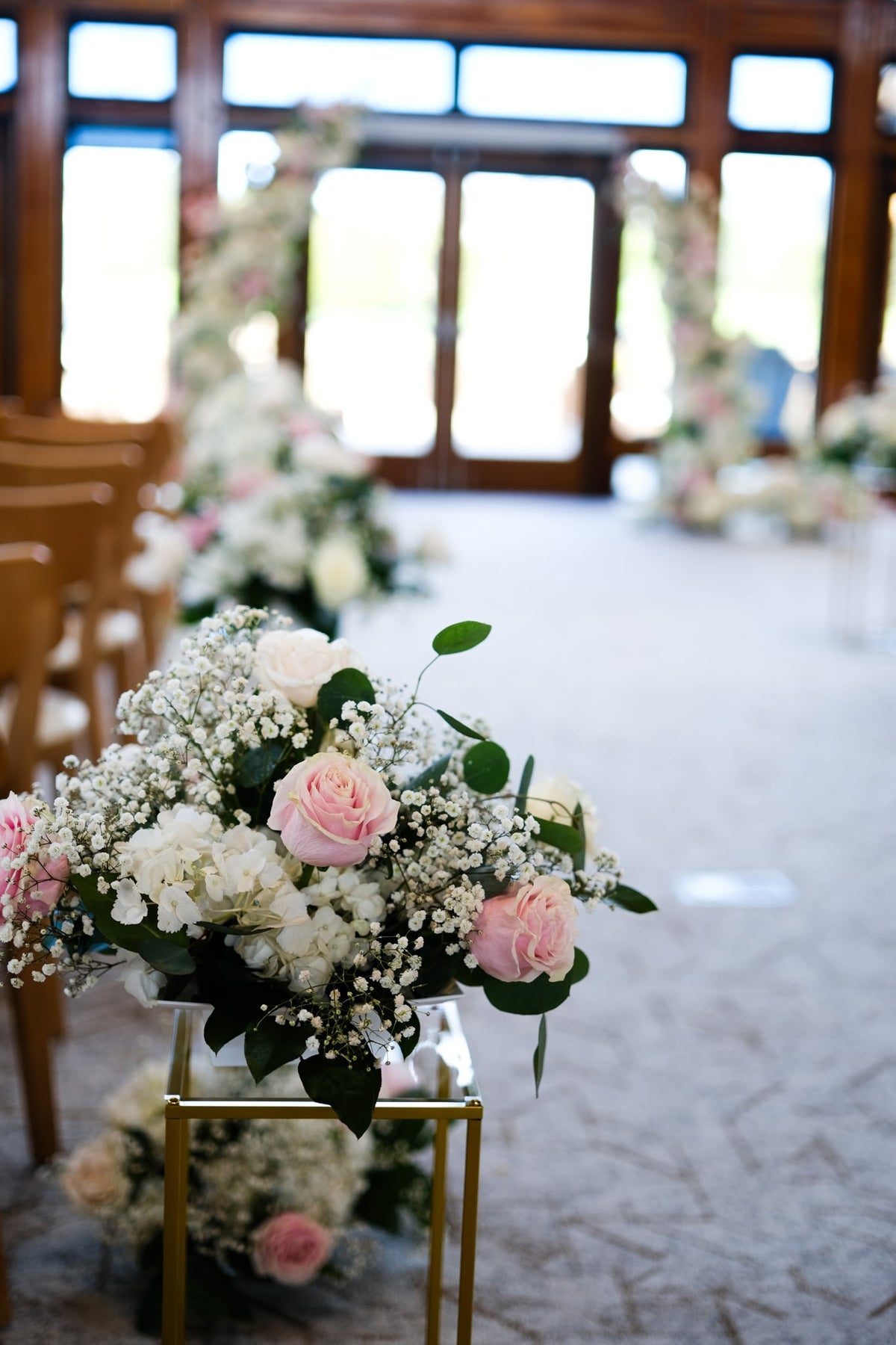 A floral arrangement of pink and white roses with baby's breath and green leaves displayed on a small gold stand in Riverway Clubhouse Burnaby, with rows of wooden chairs and bright windows in the background.