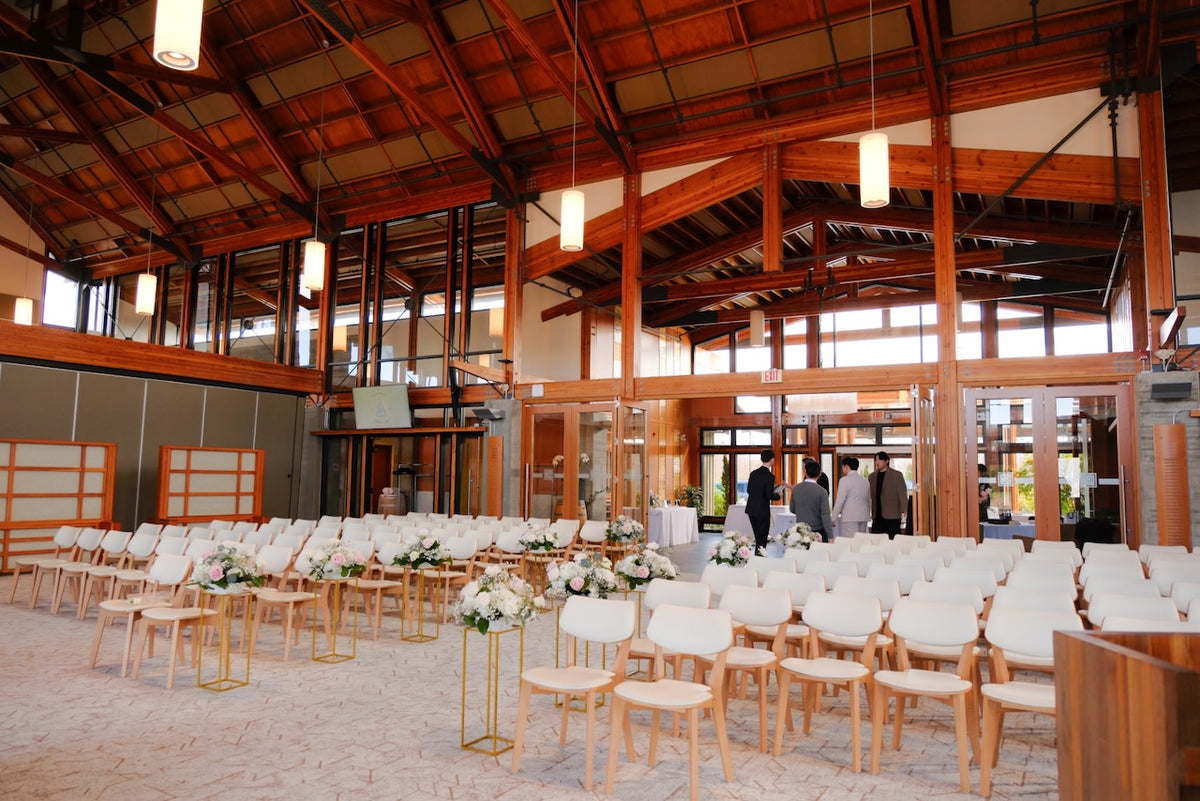 A large indoor wedding space at Riverway Clubhose Burnaby with rows of white chairs and floral arrangements, set in a spacious hall with exposed wooden beams, hanging lights, and glass doors. A few people are visible in the background near the entrance. 
