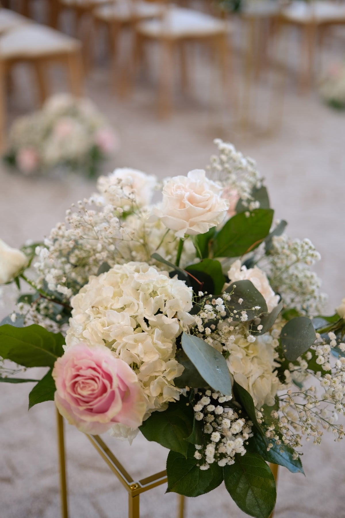 A floral arrangement featuring white and blush pink roses, white hydrangeas, baby's breath, and green foliage, displayed on a gold stand in a softly blurred event setting.