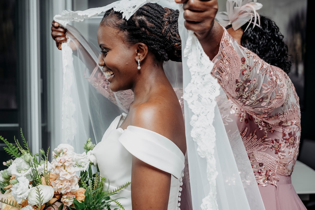 Bride with a veil being adjusted by a woman, surrounded by flowers.