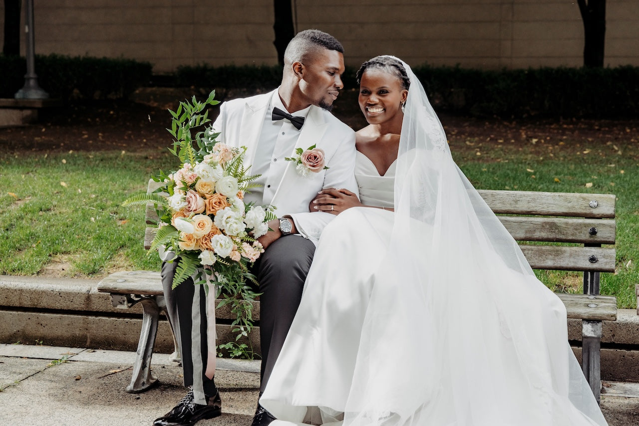 Couple in wedding attire sitting on a bench with a floral arrangement.