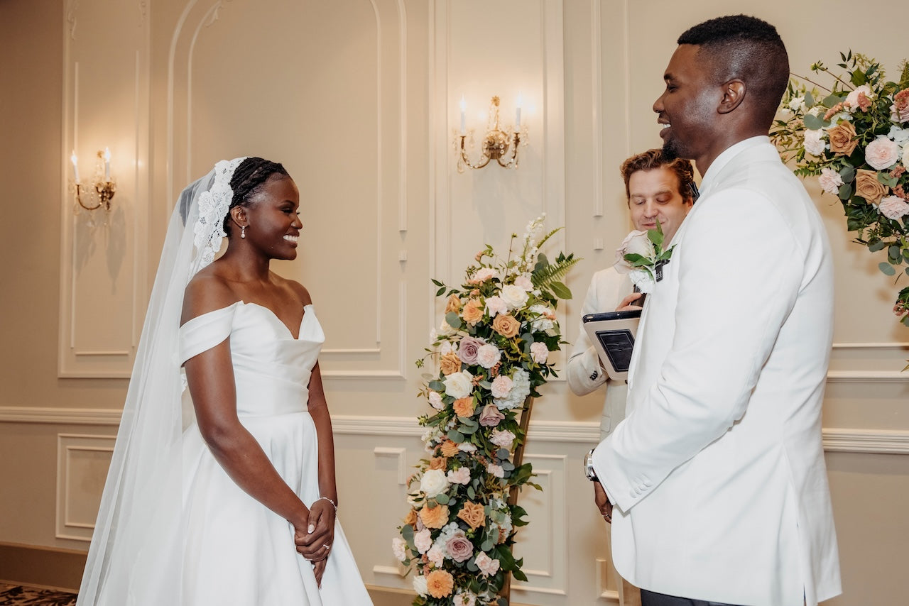 Couple in wedding attire standing in a decorated room with floral arch arrangements.