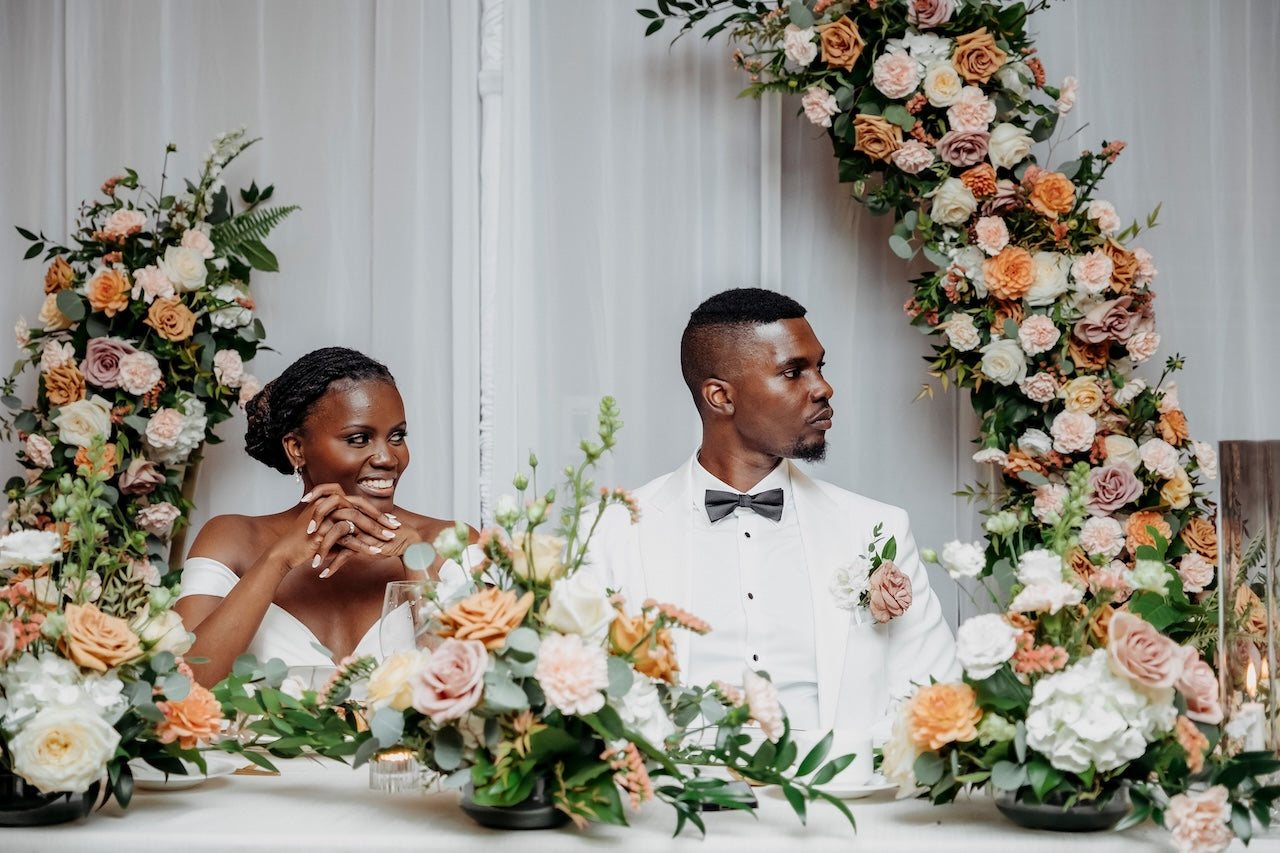  Vancouver Couple sitting at a decorated table with floral arch arrangements on either side, against a white curtain background.