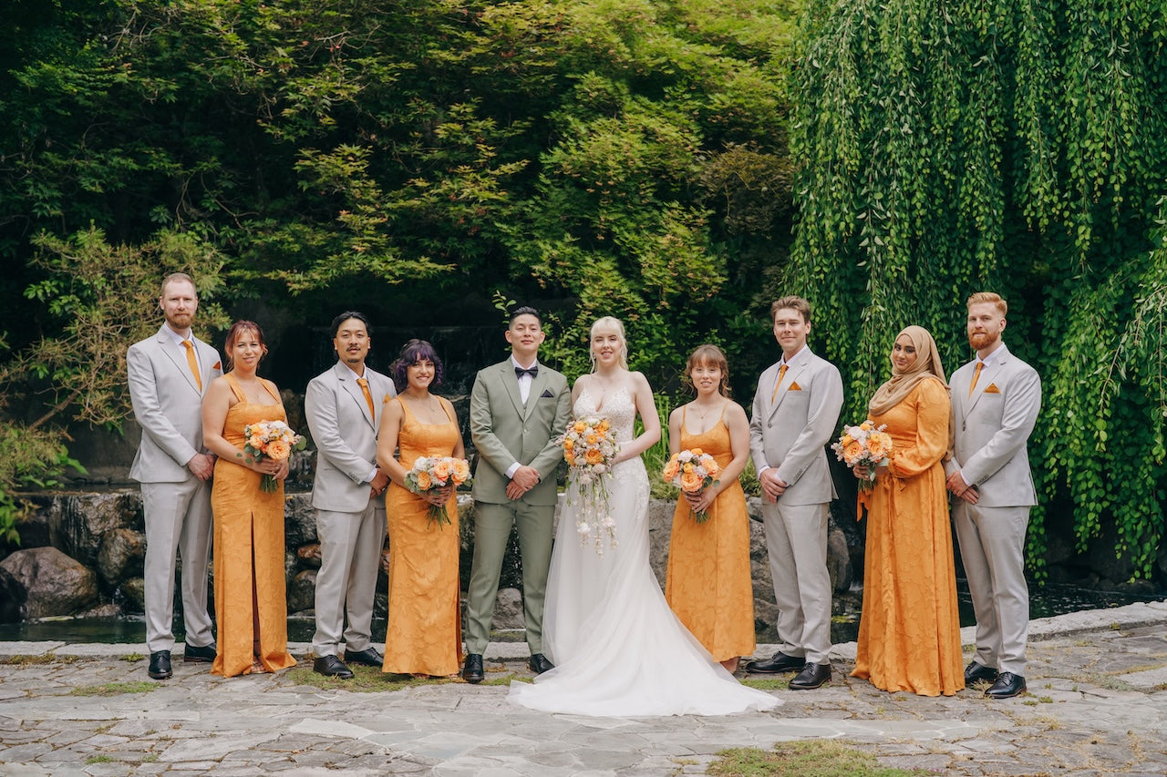 Wedding party in orange and gray standing outdoors with greenery in the background