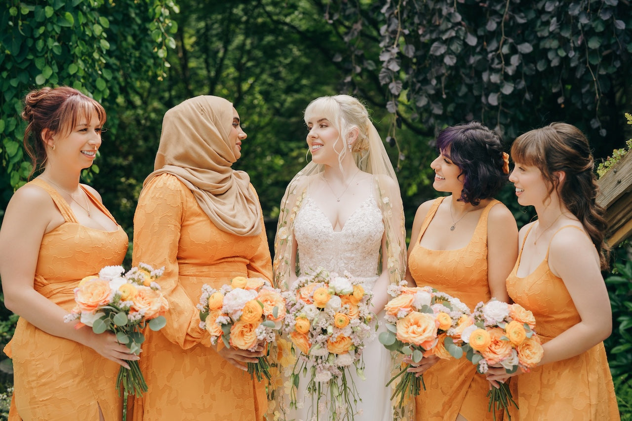 Group of women in orange dresses with floral bouquets, standing in front of green foliage.