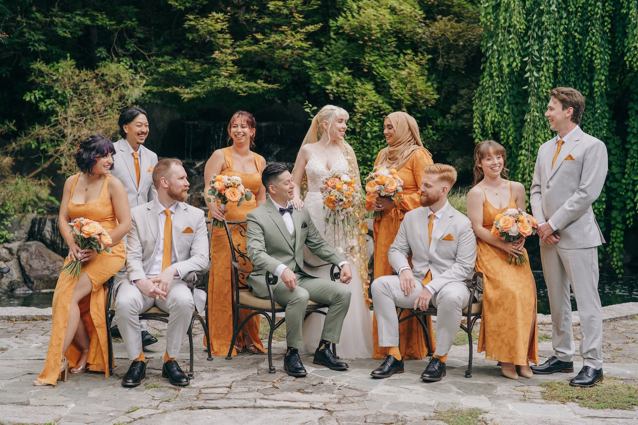 Wedding party in orange and gray attire sitting outdoors with greenery.