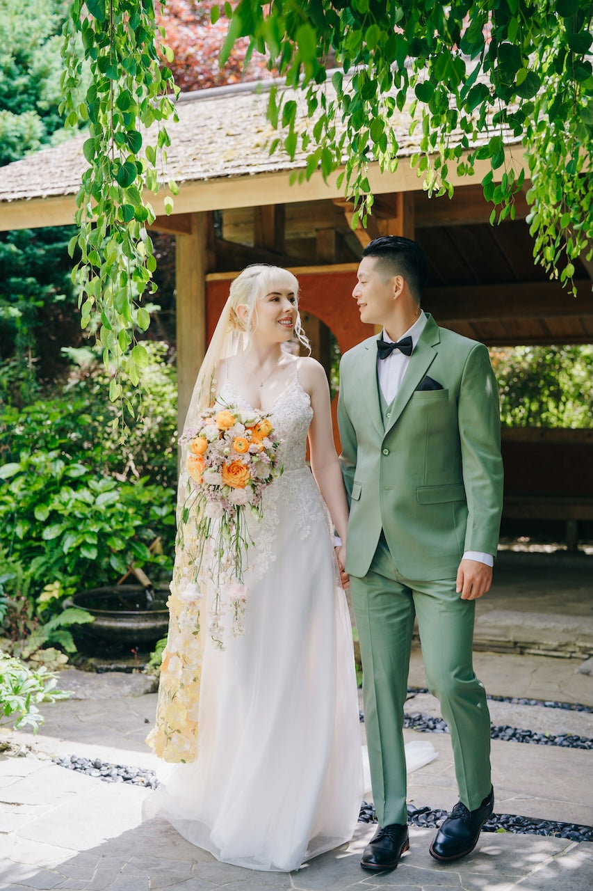 Couple in wedding attire standing outdoors with greenery around