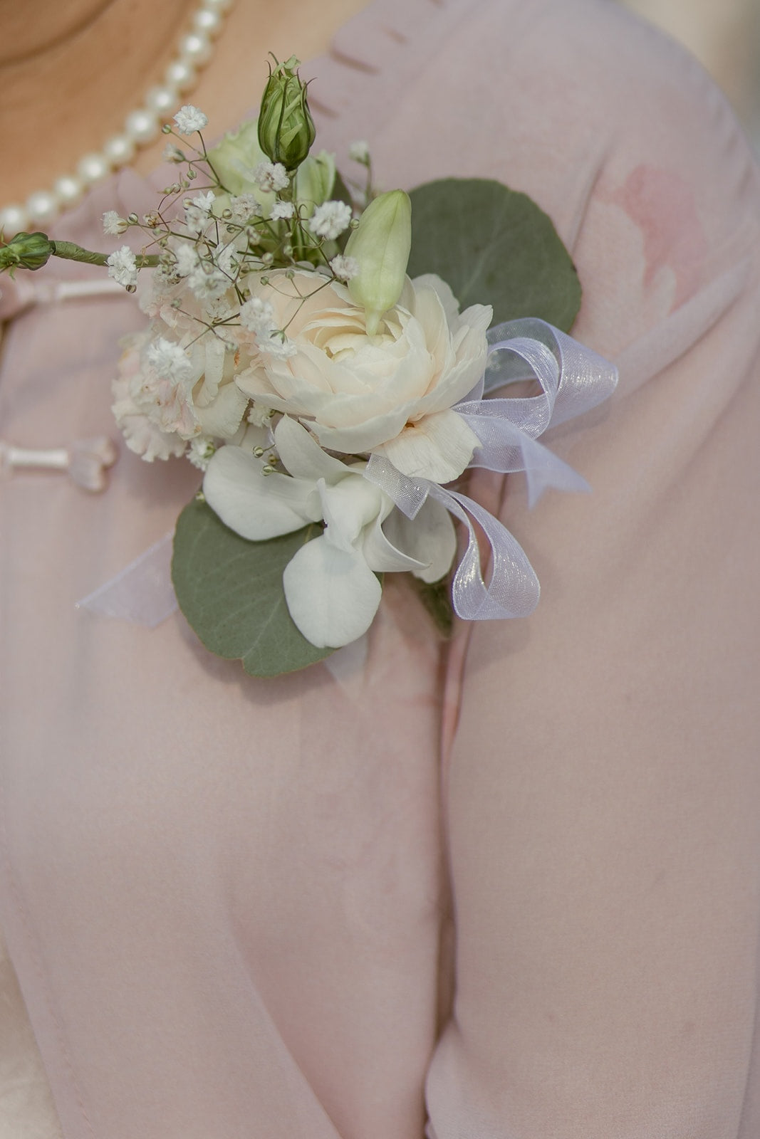 Elegant white and blush wedding boutonnière with soft floral accents and ribbon on a pale pink dress.