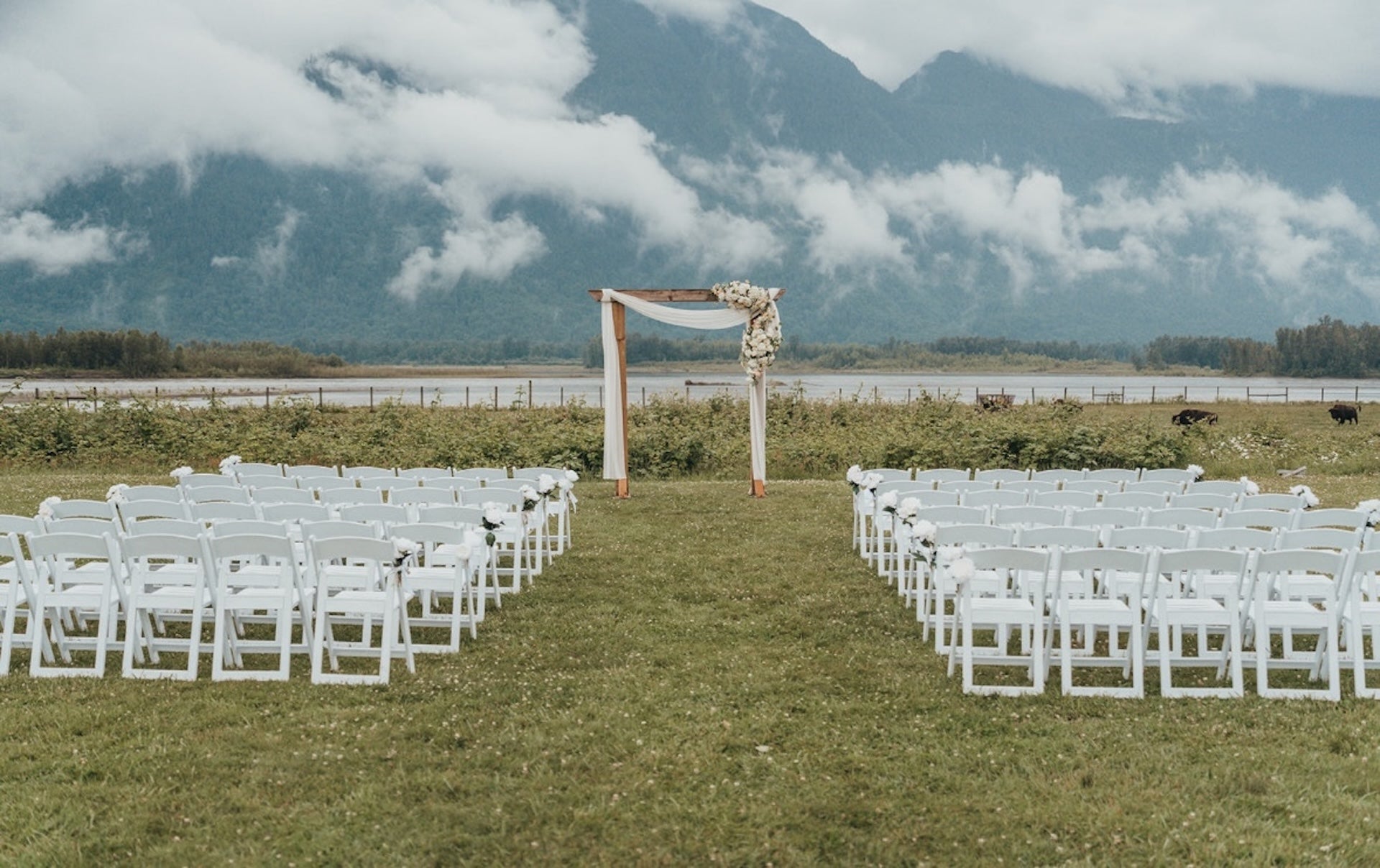 Outdoor wedding setup with chairs and floral arch against a mountainous backdrop