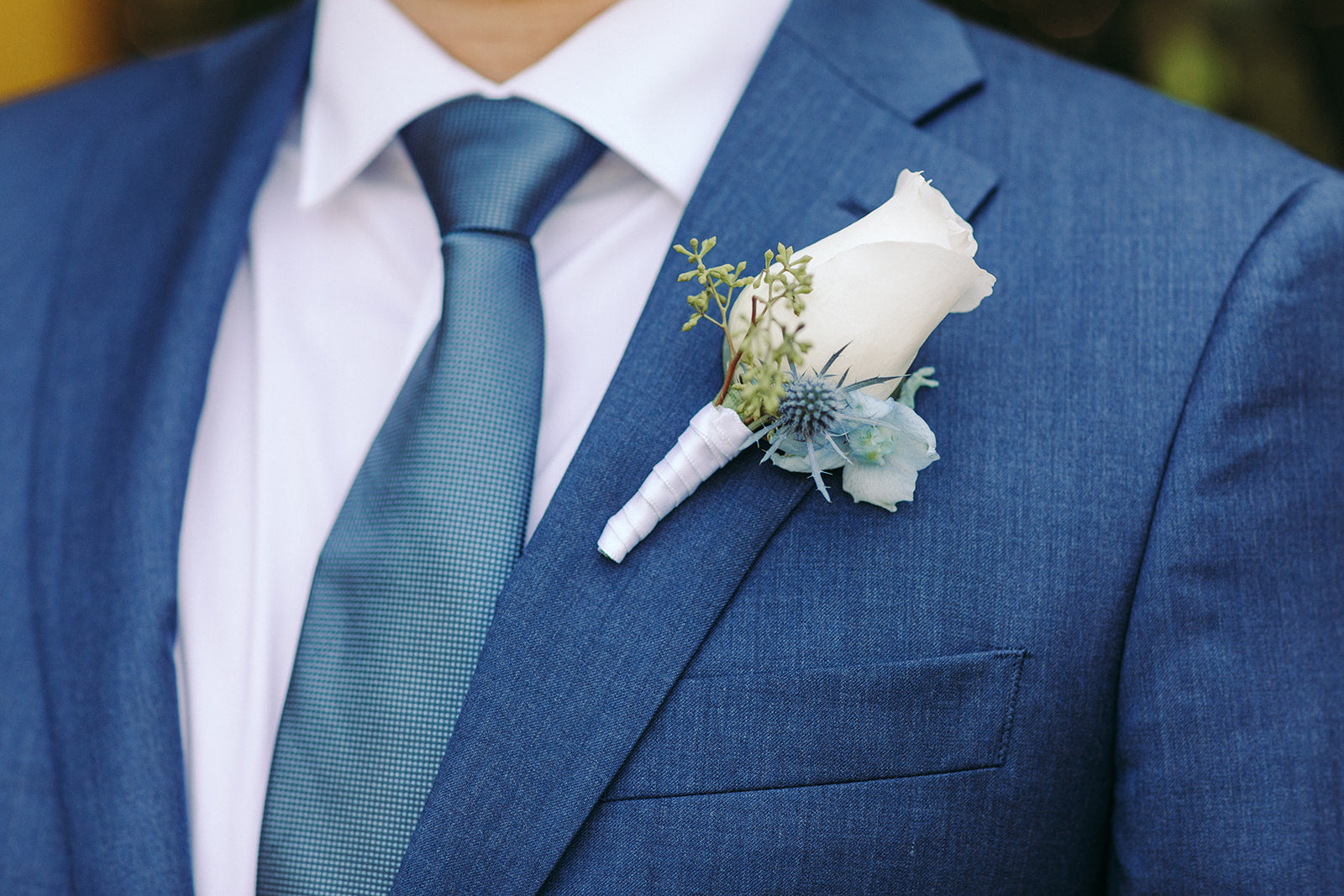Close-up of a blue suit with a white flower boutonniere and teal tie.
