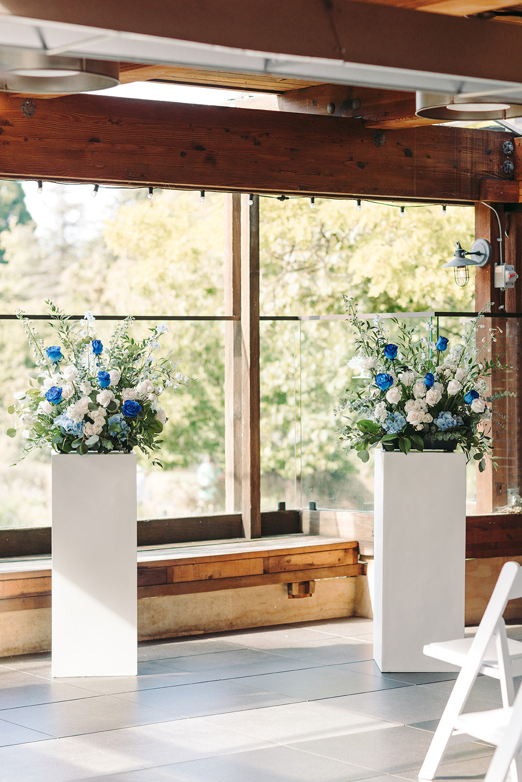 Two flower arrangements in white cylindrical pots on a wooden floor with large windows in the background.