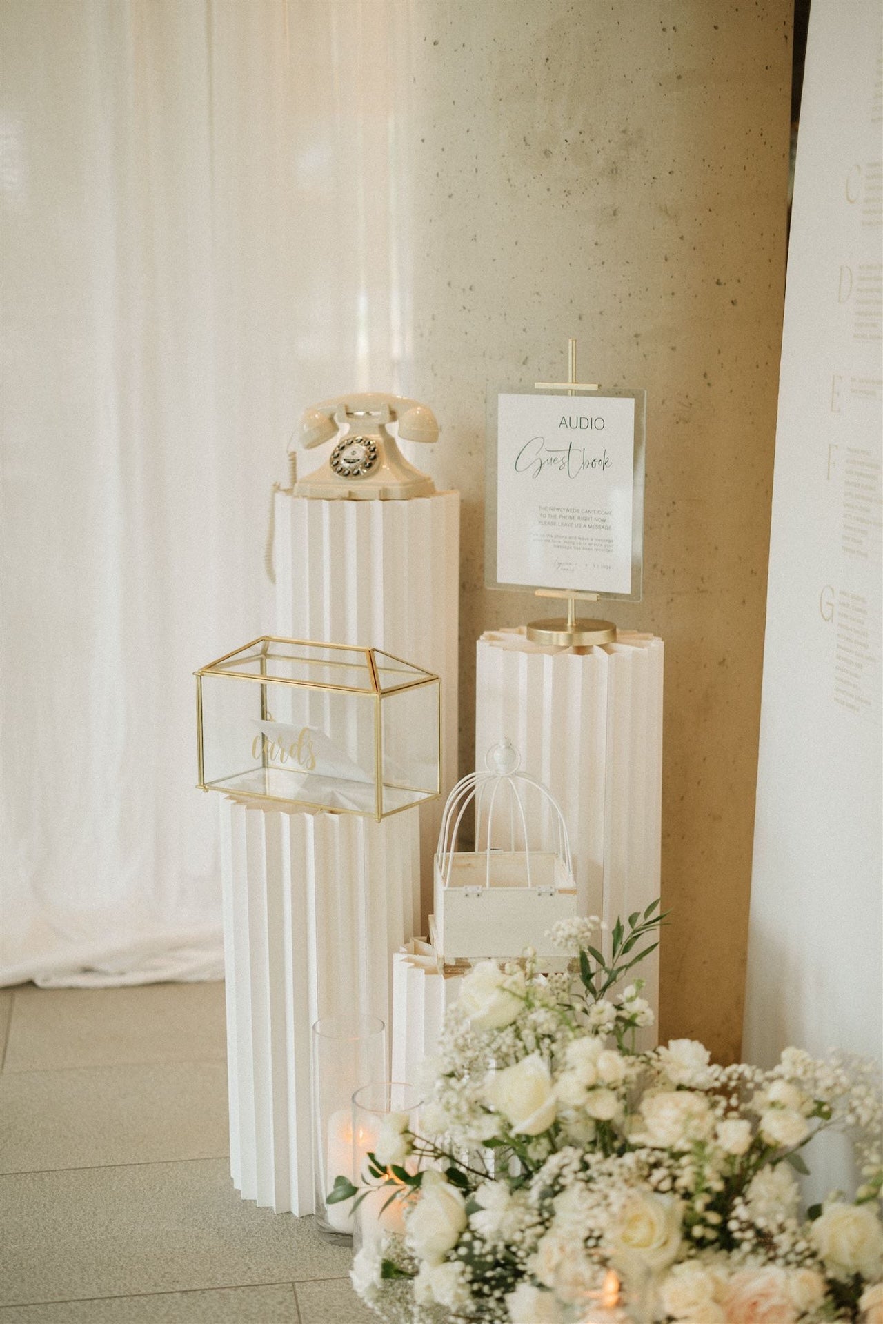 Decorative setup with flowers, a small table with a phone, and a sign on a white curtain background at a wedding reception.