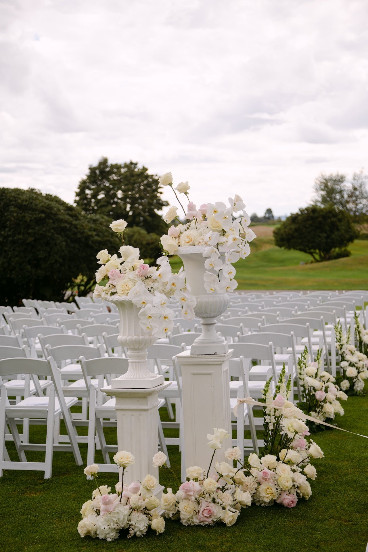Decorative floral arrangement on a white pedestal in an outdoor setting with chairs and greenery.